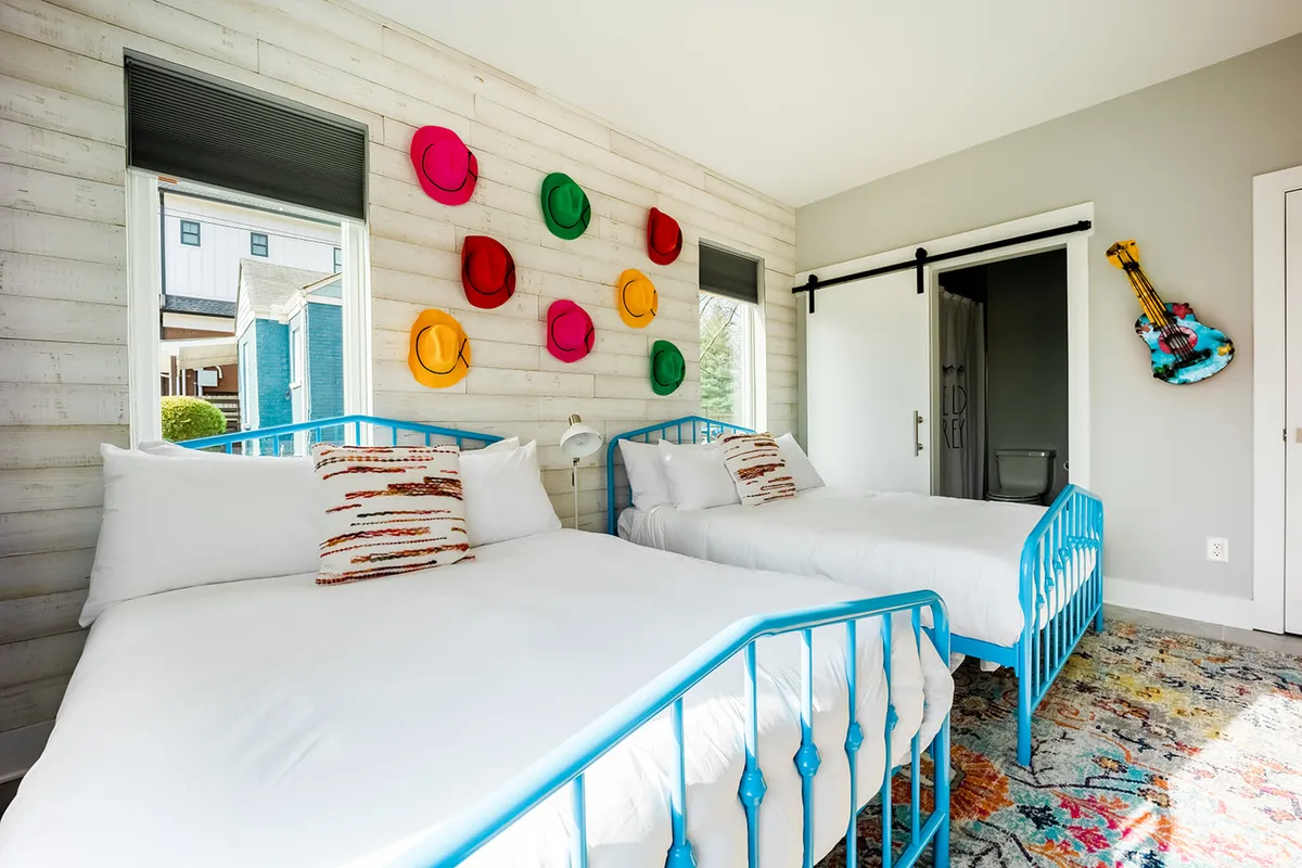 A playful bedroom with blue metal bed frames, colorful hats on the wall, and a decorative rug. The setup suggests a fun, family-friendly environment ideal for children or group stays.