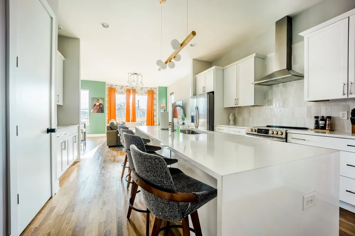 A bright and welcoming kitchen and dining area, featuring a spacious island with bar stools, large windows with orange curtains, and decorative lighting. The space is designed for both entertaining and casual dining.