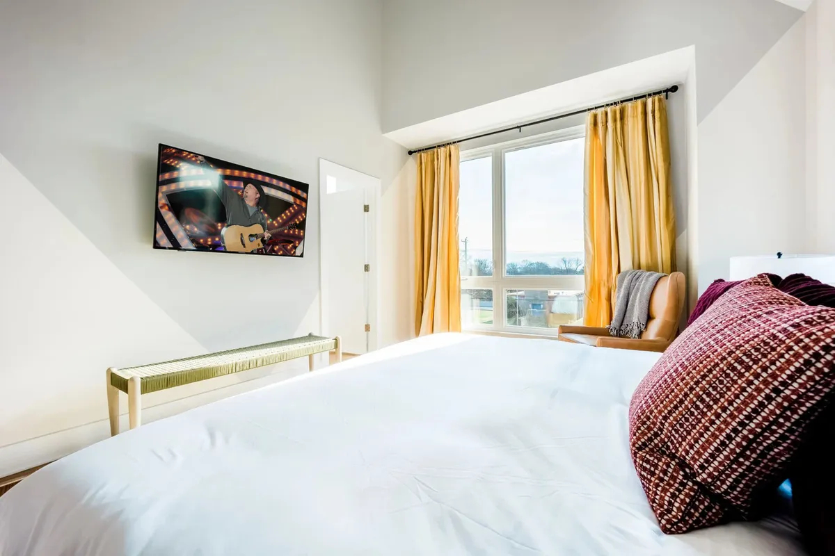 A sun-drenched bedroom featuring a plush bed dressed in soft linens, decorative pillows, a Smart TV with a performance on screen, and a large window drawing in natural light. The cozy setup exudes comfort and modern amenities ideal for relaxation.