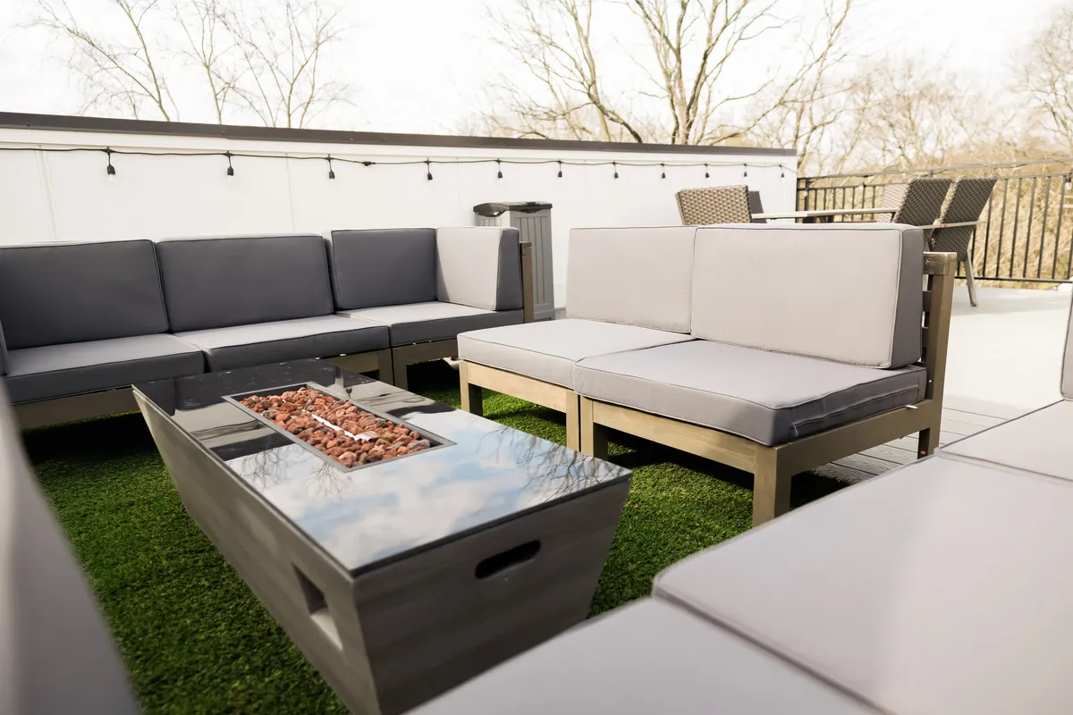 A well-appointed rooftop seating area featuring plush chairs and a fire pit, set upon artificial grass. The space is designed for both daytime relaxation and evening social gatherings, with ample room for groups to gather comfortably. The fire pit and seating arrangement create a cozy corner ideal for enjoying the outdoors year-round.