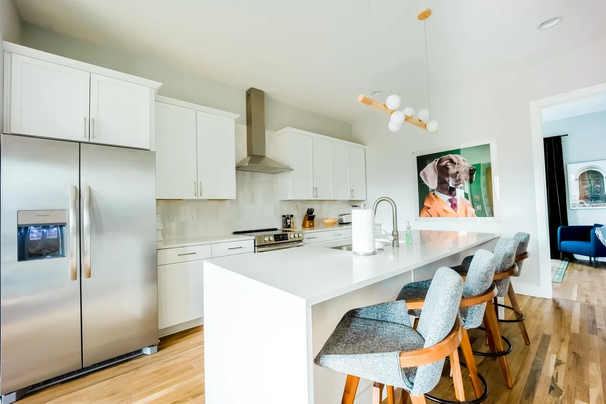 A sleek, modern kitchen with a stylish island, contemporary bar stools, stainless steel refrigerator, and playful dog portrait on the wall. The island provides ample space for meal prep or socializing.