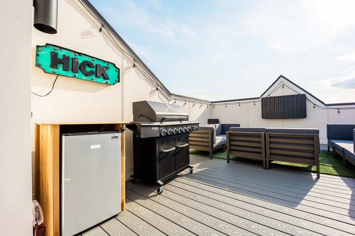 A modern rooftop terrace with a barbecue grill, generous outdoor seating, a mini refrigerator, and the quirky 'HICK' sign. There’s a sunset glow on the horizon, enhancing the sociable and relaxed atmosphere ideal for gatherings and alfresco dining.