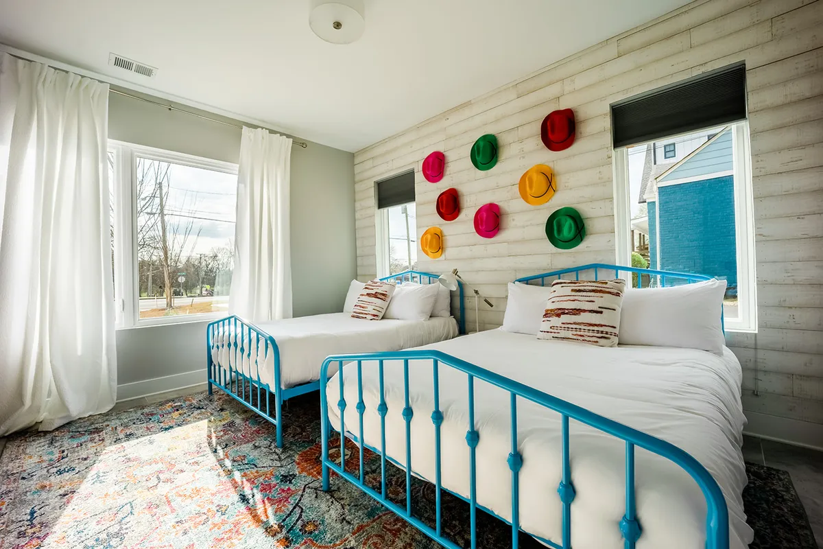 A playful and vibrant bedroom featuring two blue metal beds, colorful hats on the wall, a bright area rug, and large windows with curtains. The space feels fun, flexible, and ideal for families or groups.