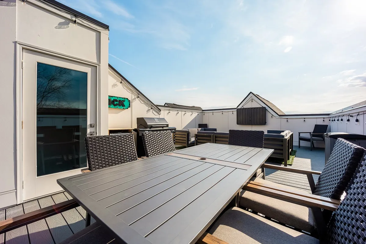 A stylish rooftop deck featuring a large outdoor dining table, comfortable chairs, a grill, and decorative signage. String lights add a festive air, and the scene is set for al fresco dining or socializing against the backdrop of the city.