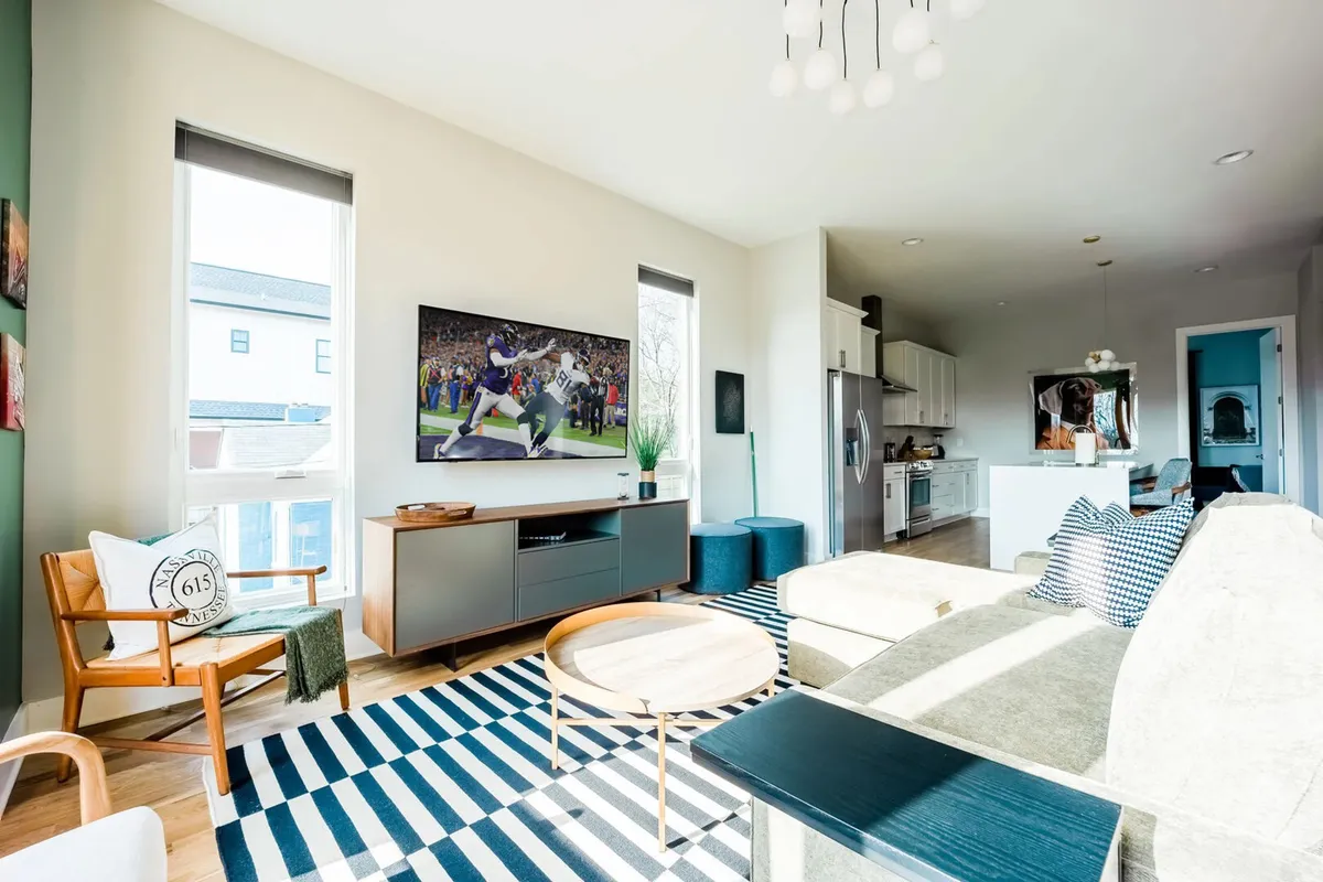 A vibrant open-concept space featuring a modern sofa and wooden accent chair arranged around a striped area rug, with a large television displaying sports. The kitchen area is visible in the background, offering a seamless flow between lounging, dining, and cooking.