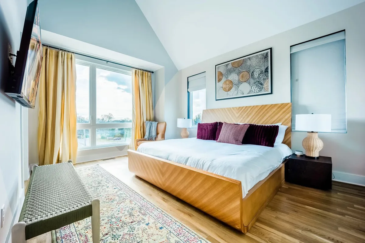 A serene bedroom scene with a large window flooding the room with natural light, a plush bed adorned with pillows, tasteful artwork, and stylish side tables, creating a comforting and restful atmosphere.
