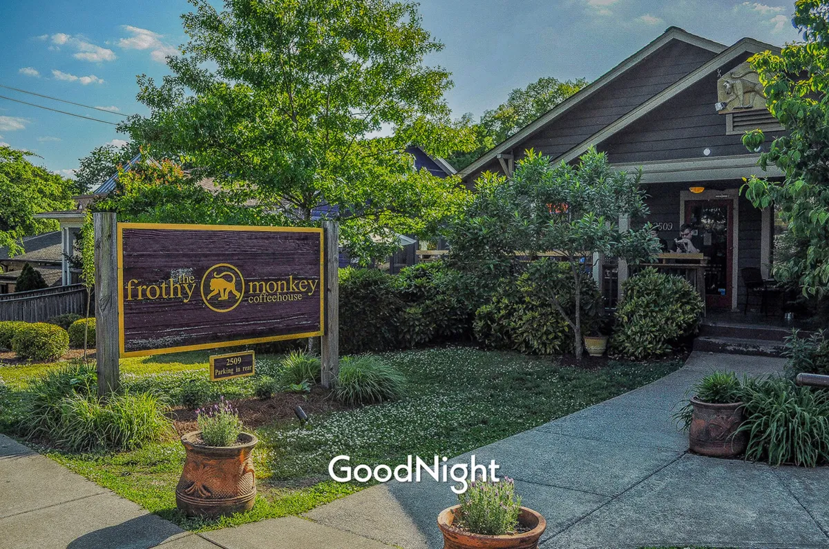 The image shows the exterior of Frothy Monkey Coffeehouse, highlighted by its sign, lush greenery, and inviting outdoor seating. The setting feels friendly and vibrant, suggesting a popular neighborhood café where locals gather, relax, and enjoy artisanal coffee.