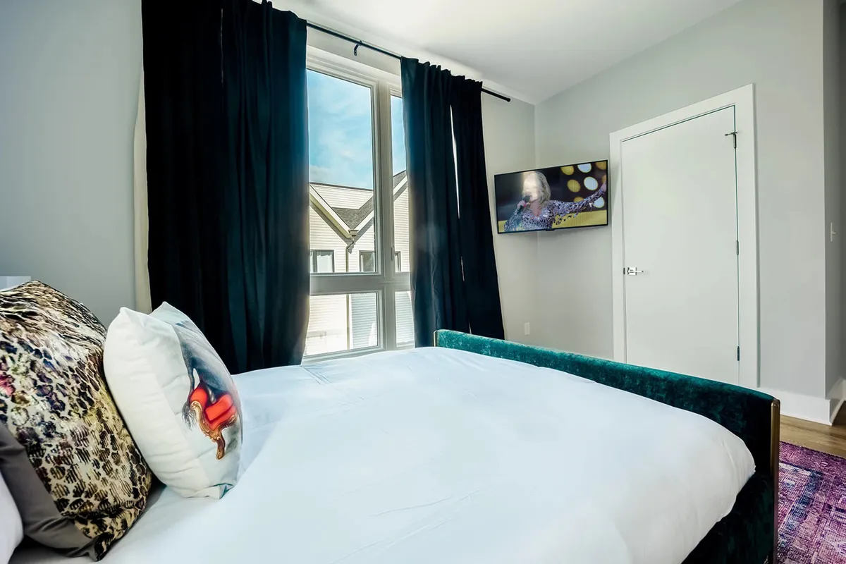 A cozy, well-lit bedroom with a bed adorned with decorative pillows, large windows letting in ample daylight, and a wall-mounted television. The overall ambiance is comfortable and inviting, suggesting a restful haven with upscale touches.