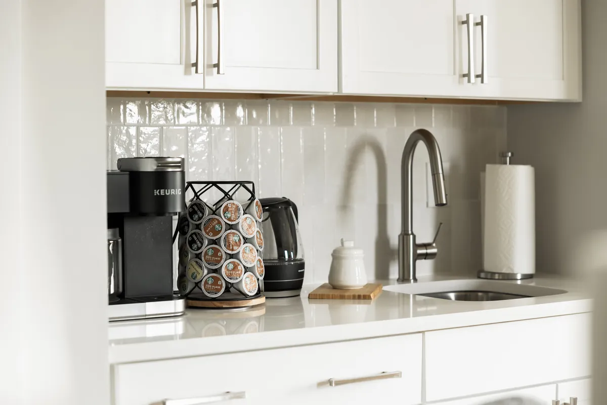 A close-up of a kitchen coffee station, featuring a Keurig coffee maker, a coffee pod holder, an electric kettle, and a sink. The area is functional and inviting, suggesting easy access to morning routines and refreshments.