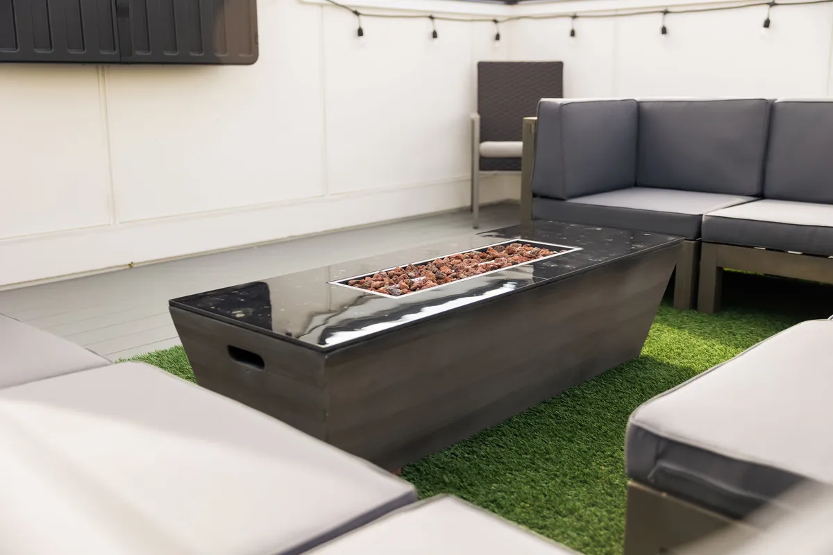 An inviting rooftop scene featuring a modern fire pit surrounded by ample seating on artificial grass. String lights overhead hint at festive nighttime gatherings, while the arrangement is perfect for sharing warm drinks and stories during cooler seasons.